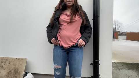 Video of a woman with long brown hair, wearing a pink shirt, black jacket, and ripped blue jeans, standing in front of a white wall and a black pipe, with a snowy outdoor scene visible through a window.