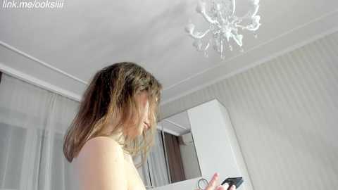 A video of a topless woman with long, wavy brown hair, looking down while holding a black object. The room has a white ceiling with a chandelier, white walls, and a mirror.