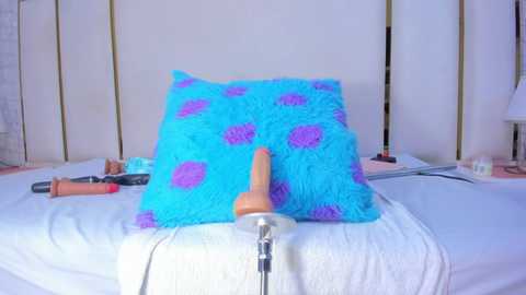 A video of a bed with a fluffy, blue pillow adorned with purple polka dots and a wooden dildo resting on it. Nearby, vibrators and sex toys lie on white sheets.