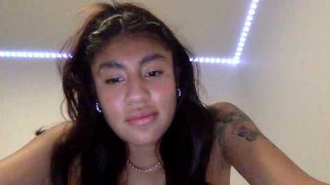 Video of a young woman with long, straight black hair, wearing a necklace, and a tattoo on her left arm, against a white wall with LED strip lights.