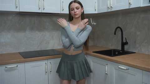 Video of a young woman with straight brown hair, wearing a grey crop top and pleated skirt, standing in a modern kitchen with white cabinets and a black sink.