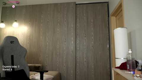 Video of a modern, minimalist bedroom featuring a gray gaming chair, beige wall, wooden sliding doors, and a white lamp on a wooden nightstand.