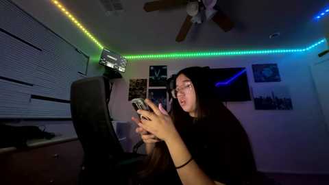 Video of a young woman with glasses, seated at a desk in a dimly-lit room with green and blue LED strip lights, playing a video game.