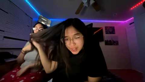 A video of two young women in a dimly lit room, one with long black hair and glasses, the other with short hair, against a backdrop of blue and pink LED strip lights.