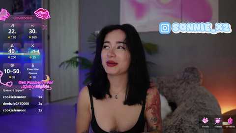 Video of a tattooed woman with shoulder-length black hair, red lipstick, and black lingerie, looking upward. Background shows a dimly-lit room with a couch, TV, and a pink floral painting.