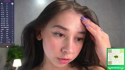 Video of a young woman with long brown hair, wearing a nose ring, touching her forehead, and with a QR code overlay. Background shows a digital clock and greenery.