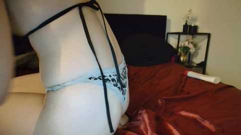 A video of a woman in a sheer black lace bra and matching panties, lying on a bed with a burgundy satin sheet. The background includes a black headboard and a small black shelf with a white orchid plant.