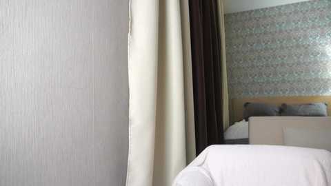 A video of a modern bedroom with light beige textured wallpaper, a bed with white sheets and a wooden headboard, and a partially visible patterned wallpaper in the background.