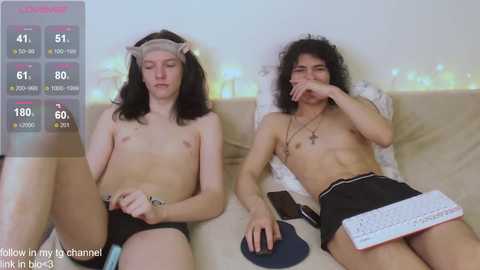 Video of two young, shirtless men with long hair, one wearing a headband, lying on a couch, one holding a phone, one with a laptop, in a cozy room.