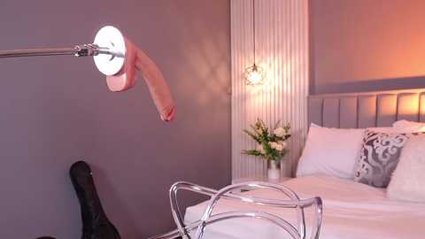 Video of a modern bedroom with a large, realistic, uncircumcised, erect penis on a silver arm attached to a wall, beside a bed with white linens, a grey pillow, and a vase of white flowers.