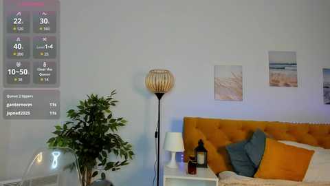 Video of a modern bedroom with a mustard-yellow tufted headboard, two pillows, and a tall floor lamp. A digital display on the wall shows climate data, and a potted plant is visible on the left.