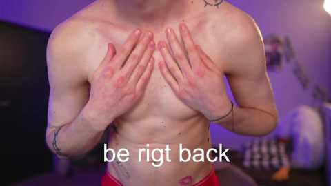 Video of a shirtless, fair-skinned man with a muscular build, covering his chest with hands, wearing red shorts, standing indoors. Background features a purple-lit room with minimalistic decor. Text reads: \"be right back.\