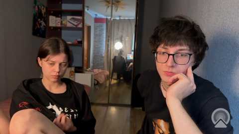 Video of two young women in a dimly lit room, one wearing glasses and a black shirt, the other with a tattoo, both looking introspective.
