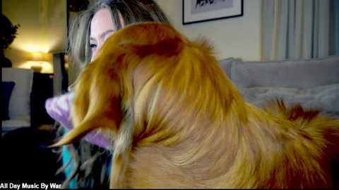 Video of a woman with long, wavy blonde hair, petting a golden retriever with a fluffy, sun-kissed coat, in a cozy living room with beige furniture and soft lighting.