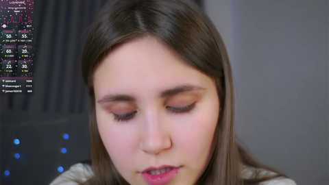 Video of a young woman with long brown hair, wearing a grey top, gazing intently at a computer monitor displaying a complex graph. The background is blurred, focusing on her serious expression and the colorful data on the screen.