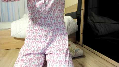 Video of a person in light pink pajamas with white and blue cartoon designs, kneeling on a light wooden floor, beside a bed with white sheets and a closed sliding door.