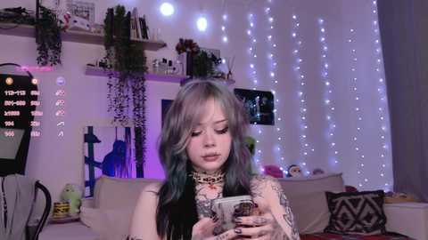 Video of a young woman with dyed hair, tattoos, and a leopard-print scarf, playing a video game, in a cozy, dimly lit room with fairy lights and decor.