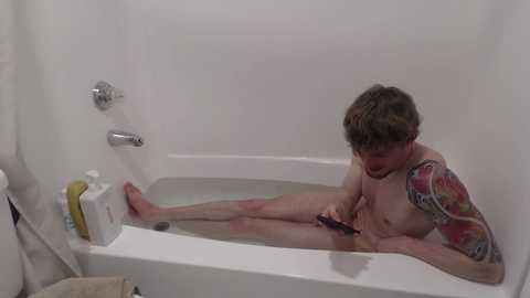 Video of a young, shirtless man with a tattoo sleeve, sitting in a white bathtub, holding a phone, water splashing around, white walls, soap dispenser, and a towel.