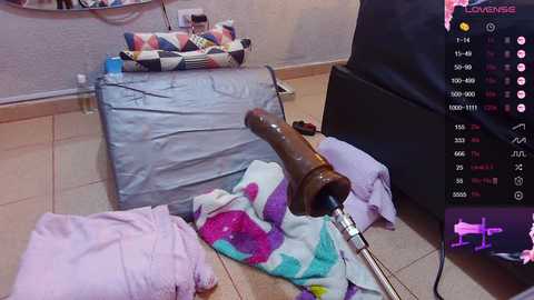 A video shows a cluttered bedroom with a large, realistic, brown dildo on a pink bedspread, surrounded by laundry, a black bag, and a colorful quilt. A live streaming overlay shows a chat window with viewers.