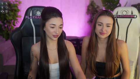 Video of two young women with long brown hair, wearing tank tops, seated in black gaming chairs with \"Playmaker\" branding, under pink lighting, in a cozy indoor setting.