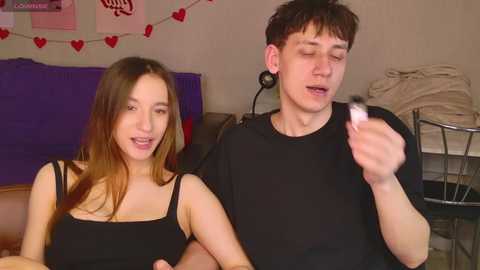 Video of a young couple indoors; the woman, with long brown hair, wears a black top, smiling. The man, with short dark hair, holds a makeup brush, wearing a black shirt. Background includes purple and white decor.