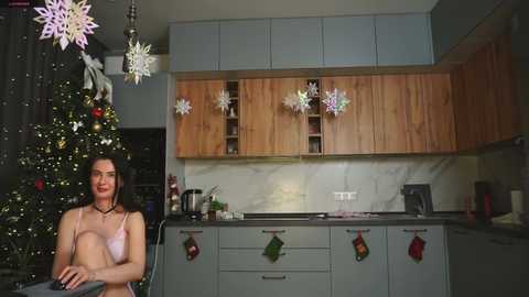Video of a woman in a pink bra and black necklace seated in a modern kitchen with wooden cabinets, Christmas decorations, and a lit Christmas tree.
