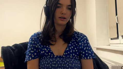 Media: Video of a young woman with light brown skin, wearing a blue dress with white polka dots, sitting on a black couch, listening to headphones, in a plain, well-lit room.