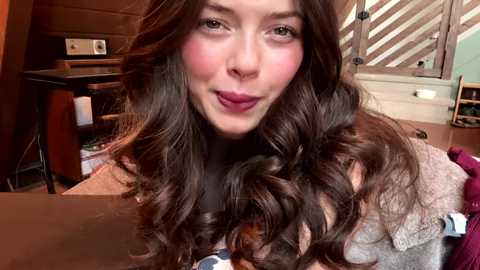 Video of a young woman with long, wavy brown hair, light skin, and pink lipstick, smiling in a cozy kitchen with wooden cabinets, windows, and a table.