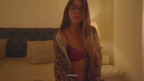 Video of a young woman with long brown hair, wearing glasses, a red lace bra, and a leopard print robe, sitting on a bed with beige linens and a gray headboard.