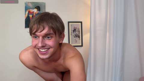 Video of a young, shirtless, Caucasian man with short brown hair, smiling widely. He is leaning forward in a bedroom with a framed painting on the wall and a curtain partially drawn.