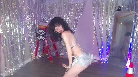 A video of a slender Asian woman with long black hair and glasses, wearing a glittery silver bikini, posing provocatively in a mirrored, shiny, silver-lined room with a dartboard and colorful lights.
