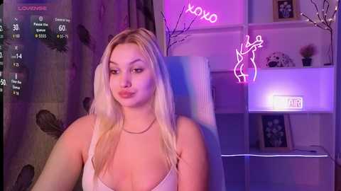 A video of a young woman with long blonde hair, wearing a white tank top, sitting in a dimly lit room with neon signs and a \"LOVE\" sign.