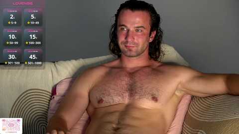 Video of a shirtless, muscular, Caucasian man with long, wavy brown hair, reclining on a beige couch. A fitness app screen in the upper left displays his stats: 2.9 BMI, 10% body fat, 190 lbs.