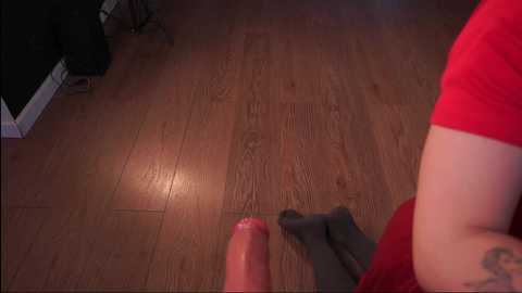 Media: A video showing a pair of legs in red and gray socks standing on a wooden floor. The legs are positioned in the foreground, with a large, pink dildo placed between them. The background includes a black piece of furniture and a beige carpet.
