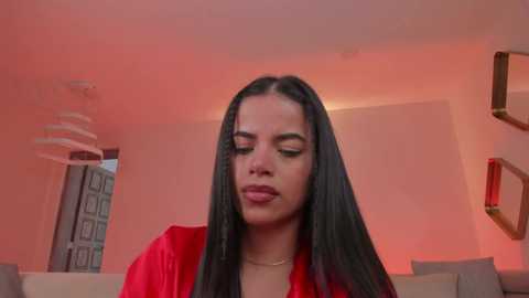 Video of a Latina woman with long, straight black hair and light skin, wearing a red dress, in a modern living room with red lighting, framed art on the wall, and a white chandelier.