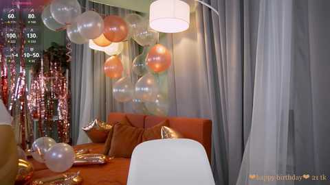Video of a modern, dimly lit room decorated for a birthday party with silver and gold balloons, sheer curtains, and orange couch.