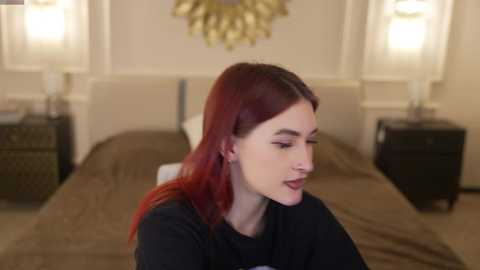 Video of a young woman with fair skin and red hair, wearing a black top, smiling in a well-lit bedroom with beige walls, white crown molding, and two bedside lamps.