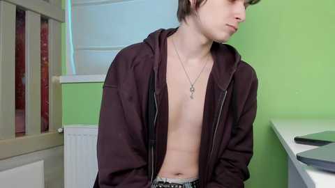 Video of a pale, skinny young man with brown hair, wearing a black hoodie unzipped to reveal his bare chest, standing in a room with green walls and white radiator.