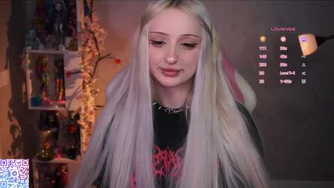 Video of a young woman with long platinum blonde hair, wearing a black shirt with red graphics, in a cozy room with shelves filled with books and decorative items.