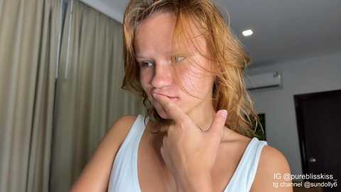 Video of a young woman with light skin and messy, wavy, shoulder-length blonde hair, wearing a light blue tank top, with her index finger to her lips in a thoughtful pose. Background features beige curtains and a dark door.