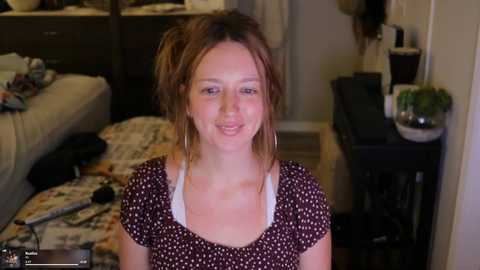 Media: Video of a young, fair-skinned woman with auburn hair in a messy bun, wearing a black polka-dot top, smiling in a cluttered living room with a white couch, potted plants, and scattered items.