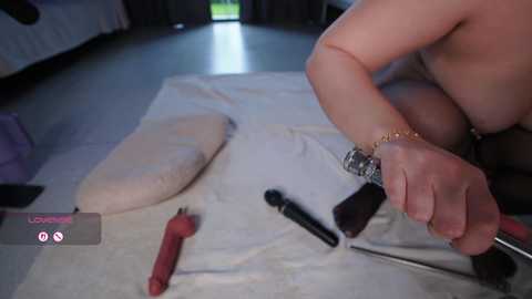 A video of a nude woman with fair skin, medium-sized breasts, and a gold bracelet, using a metal tool on a white towel spread on a dark floor. Various sex toys are scattered around, including a vibrator.