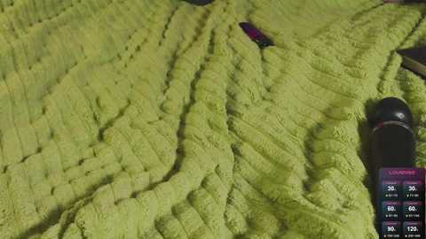 Video of a green, textured, quilted blanket with a black camera lens on the right side, showing a weather forecast display with temperature readings in the upper right corner.