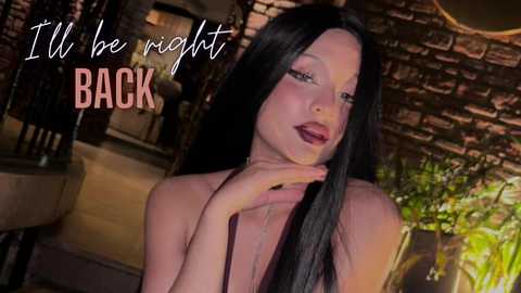 Video of a fair-skinned woman with long black hair, wearing a sleeveless top, gazing pensively. Background features a rustic brick wall and green foliage. Text reads, \"I'll be right back.\