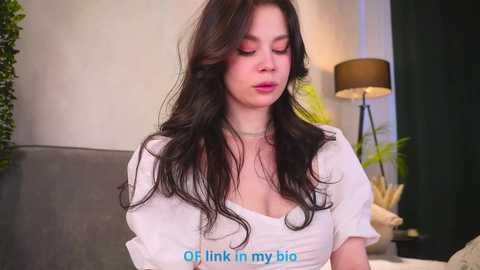 Video of a young, fair-skinned woman with long, wavy black hair, wearing a white top, standing indoors. Text overlay reads, \"Of course I link in my big.\