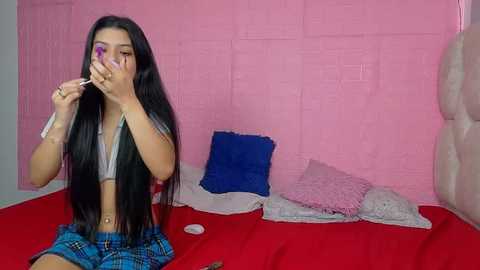 A video of a young woman with long black hair, wearing a gray crop top and blue plaid skirt, applying makeup while sitting on a red bed with pink and blue pillows against a pink brick wall.