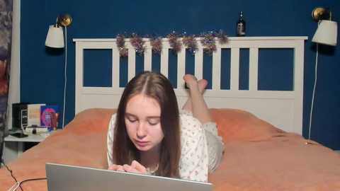 Video of a young woman with long brown hair, wearing a white floral nightgown, lying on an orange bedspread, focusing on a laptop, in a blue-painted room with a white wooden headboard.