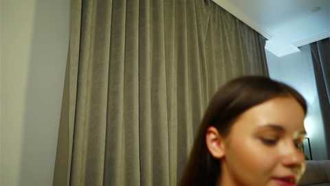 A video of a woman with long brown hair, fair skin, and closed eyes, facing right. She stands in a modern room with gray curtains and white walls, partially visible behind her.