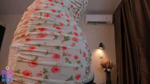 Video of a woman in a tight, floral-patterned dress, accentuating her curves, standing in a dimly lit room with beige walls, a brown curtain, and a small decorative vase.