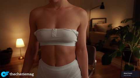 Video of a slim, fair-skinned woman with a toned midriff, wearing a strapless white bandeau top and matching high-waisted shorts, standing in a dimly-lit living room with a lamp, plants, and framed pictures on the walls.